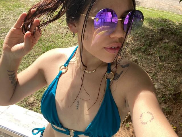 SophiaMarx in a blue bikini, showing tits with small tan lines, wearing purple mirrored sunglasses, holding wet brown hair, tattoos on arms, and a pearl necklace. Sunlit outdoor.