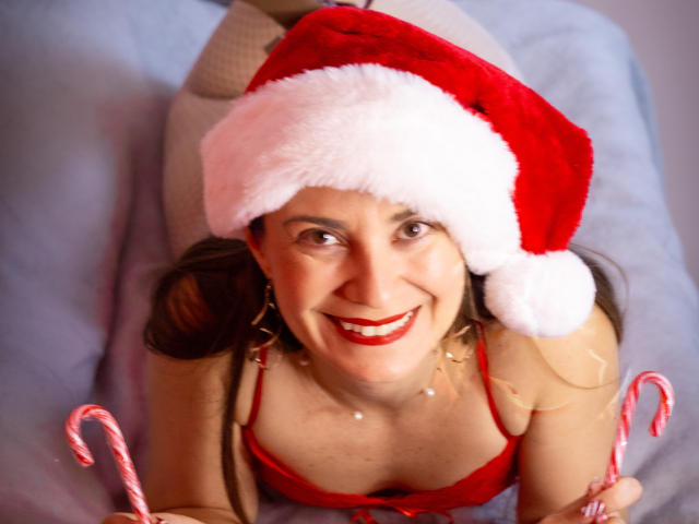 NatashaKute with red Santa hat, red bra, holding candy canes, smiling, showing her tits, wearing white socks, lying on bed, red lipstick, pearls around neck.