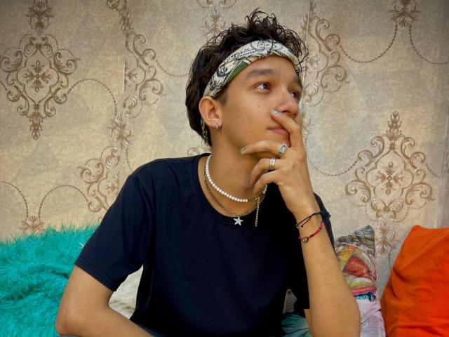 KristofMills with short curly hair, wearing a bandana, black tee, pearl necklace, and ring, thinking with hand on chin, green nails, and a colorful pillow behind.