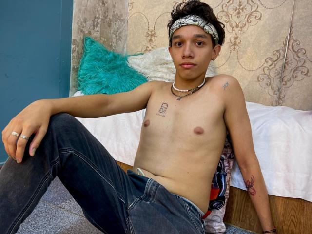Shirtless KristofMills with a small chest, wearing a headband, jeans, and a necklace, sitting on a bed with a turquoise pillow, tattoos on chest and arm, looking serious.
