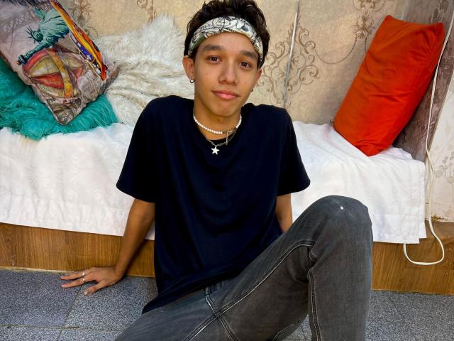 KristofMills is a cute young dude with short brown hair, wearing a black shirt, grey jeans, and a white headband, sitting on a bed with colorful pillows.
