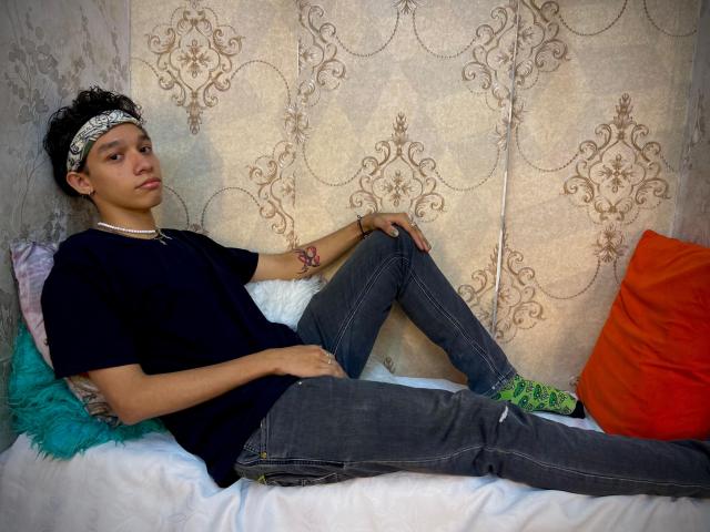 KristofMills with curly black hair, wearing a black tee, jeans, green socks, headband, and necklace, lounging on a bed with teal, orange, and pink pillows.