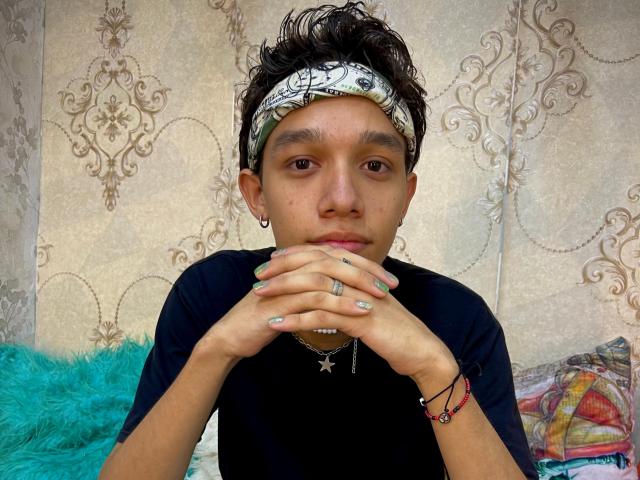 KristofMills with a headband, black shirt, and ring, hands over mouth, green nail polish, and star necklace, looking serious. Hands are on turquoise pillow.