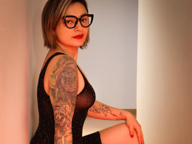 KatParadise with glasses, short blonde hair, red lipstick, black sheer dress, showing a tiger tattoo on her arm, sitting, looking at camera, right hand on knee, sexy.