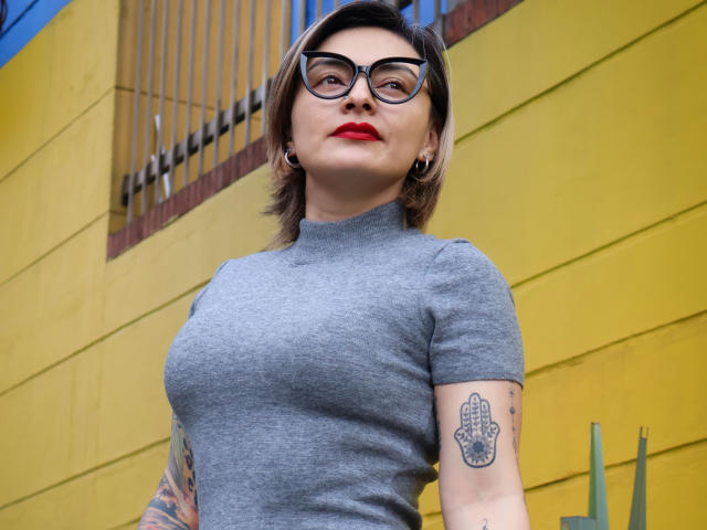 KatParadise avec des lunettes, du rouge à lèvres rouge, des cheveux blonds courts, un t-shirt gris, des tatouages sur les bras, regardant vers le bas, un fond de mur jaune, de petites boucles d'oreilles, une expression confiante, un sourire subtil..