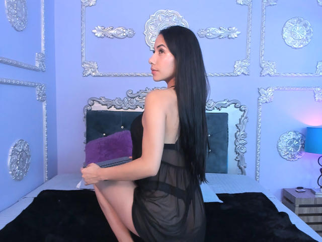 ArianaRogers with long black hair, wearing a sheer black dress, sitting on a bed, holding a purple pillow, showing her ass, with a laptop in her hands.
