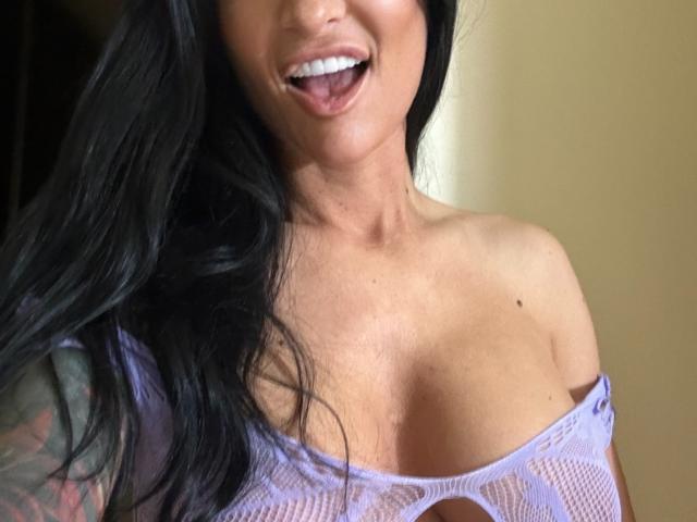 AndraD with black hair, smiling with tongue out, wearing a purple lace top that reveals her big boobs, showing a tattoo on her left arm.