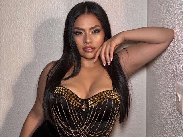 BellaSmithX69 with long black hair, wearing a black dress with gold chain details, large tits, and a serious expression, leaning against a white wall with one hand up.