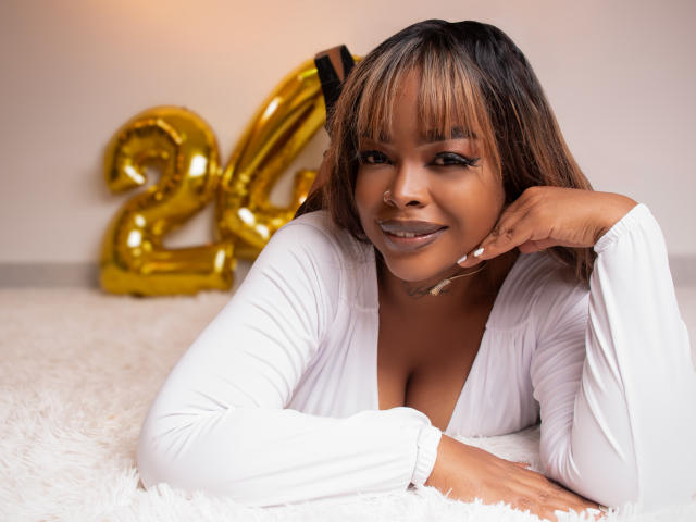 MadinsonLopez, with brown skin and blonde highlights, laying on a white fluffy rug, wearing a white, deep V-neck, smiling at the camera, with a gold "24" balloon behind her.