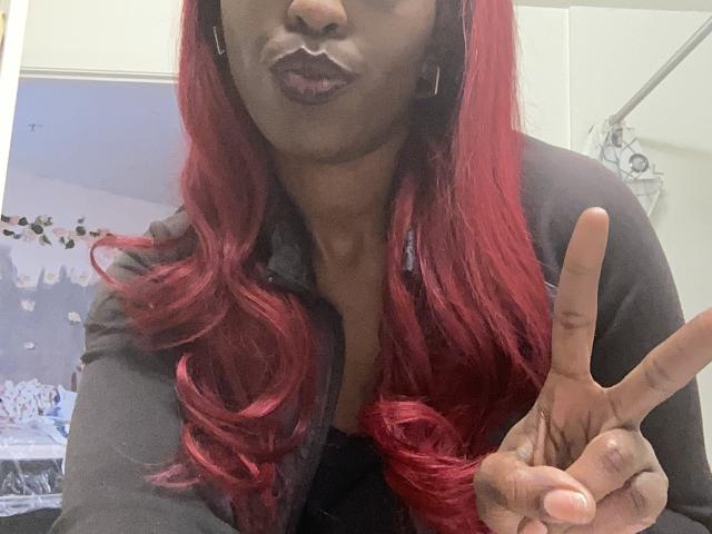 BellaStella69 with red hair, dark lipstick, black shirt, and gold earrings, making a peace sign, with a small amount of white fluid on her lips.