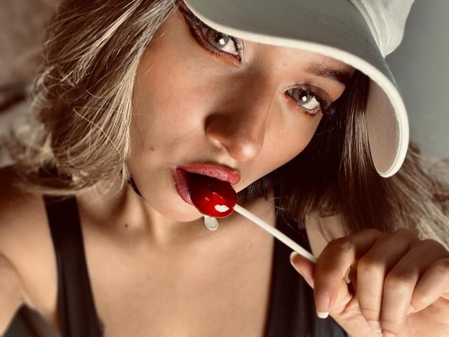 LillyTaylor with light brown hair, wearing a white cap, black tank top, and red lipstick, sucking on a red lollipop, holding it with her right hand.