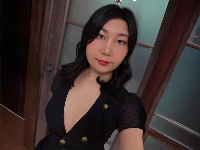 SoftGeraldine, avec ses cheveux noirs mi-longs, son rouge à lèvres rose et sa robe noire transparente ornée de boutons dorés, pose en selfie, dévoilant sa poitrine. Elle regarde l'appareil photo avec un léger sourire..