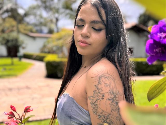 KarinaMenez, con lunghi capelli scuri, gli occhi chiusi, indossa un top blu senza spalline, mostrando un tatuaggio di un sole con una donna sul braccio destro, in piedi vicino a fiori colorati..
