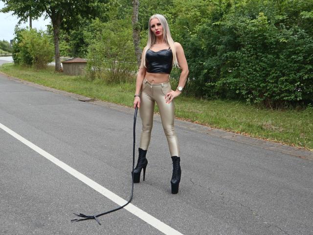 Miss-Mila with blonde hair, black leather crop top, gold shiny pants, black high heels, holding black whip, standing on empty road, green trees in background.