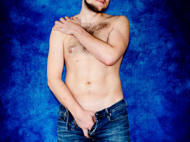 Shirtless OrgasmMaker with a hairy chest, blue jeans, hand covering his flaccid, uncircumcised penis, and a beard, standing against a blue textured wall.