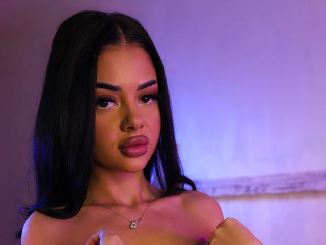 MolyStar with long black hair, full eyebrows, and pink lipstick, showing off her small, perky tits with a nose ring and a necklace, under purple and blue lighting.