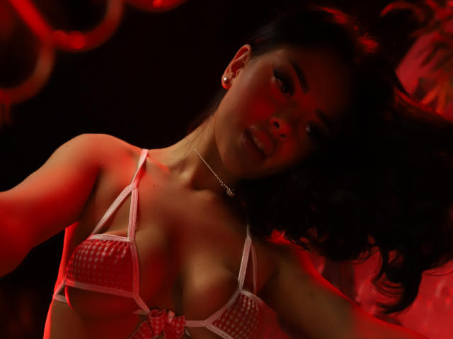 MolyStar with red-lit skin, wearing a pink checkered bra that barely covers her tits, has dark hair, and a small necklace, looking up with a slight smile.