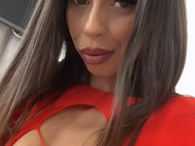 CandieCrossLand with long brown hair, full red lips, and wearing a red top with a cutout, showing a hint of her tits. Her eyes are slightly upturned.