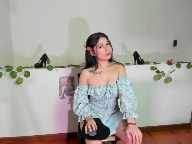 Hesttia mit schwarzen Haaren, einem roten Blumen in den Haaren, einem blauen Kleid mit Schulterpartie, schwarzen High Heels, einem Tattoo auf der Brust, sitzend auf einem Stuhl, Holzboden, grüne Blattdekorationen, leicht schmollender Gesichtsausdruck..