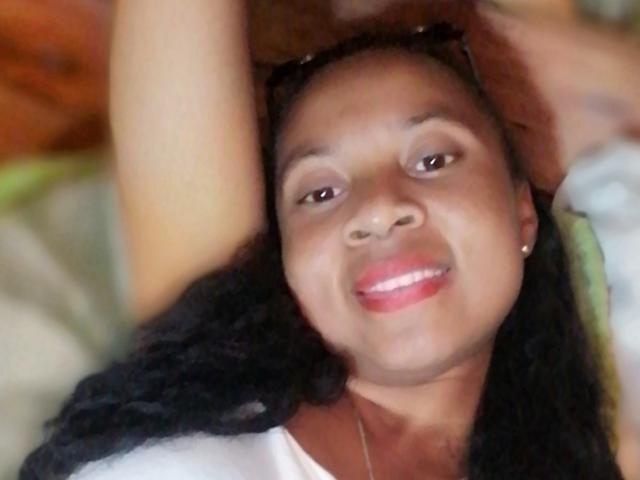 Edrwina69 has dark brown skin, black curly hair, and is smiling with pink lipstick, wearing a white shirt, with her arm raised, and sunglasses on her head.