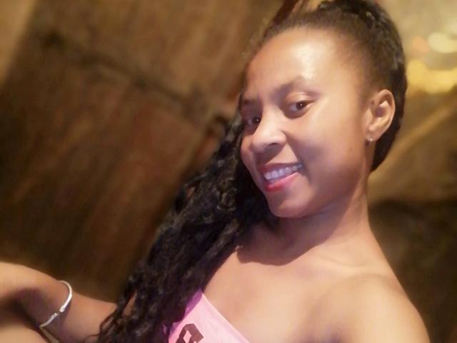 Edrwina69 with dark skin, long braided hair, smiling, showing teeth, pink top, bracelet, and bare shoulders, in a warm-lit setting.