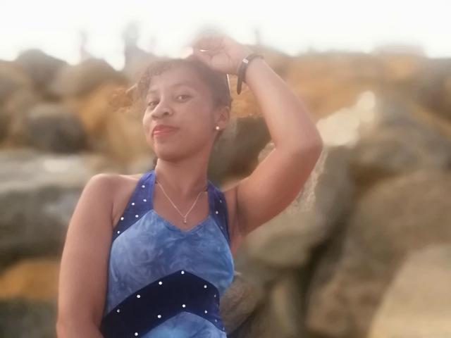 Edrwina69 with short curly hair, wearing a blue halter dress with white polka dots, raising her right arm, smiling, sunlight in the background, looking slightly to the side.