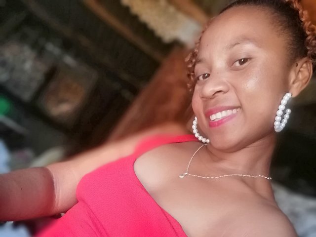 Edrwina69 with dark brown skin, curly red hair, wearing a red top, pearl earrings, and a necklace, smiling at the camera, showing her small tits and pink lipstick.