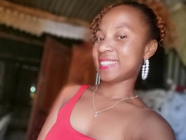 Edrwina69 with dark skin, braided hair, red tank top, pink lipstick, smiling, showing teeth, wearing dangling earrings and silver necklace, medium tits, confident, indoors.