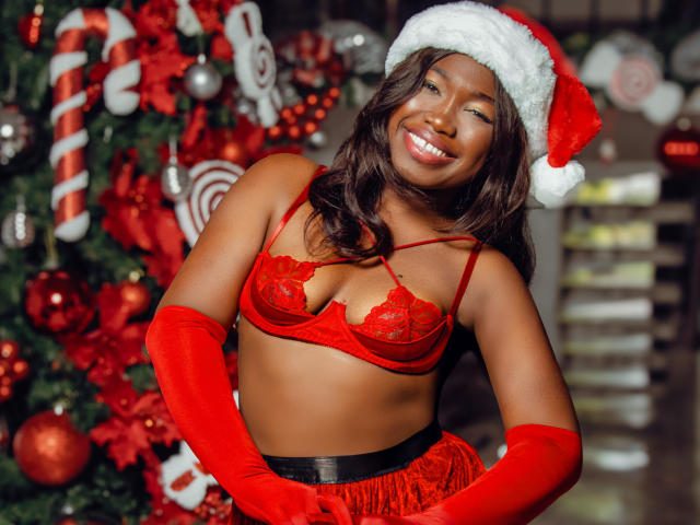 KarlieMoss with dark skin, wearing red lace bra and panties, red gloves, Santa hat, smiling, showing her tits, standing in front of a Christmas tree with red decorations.