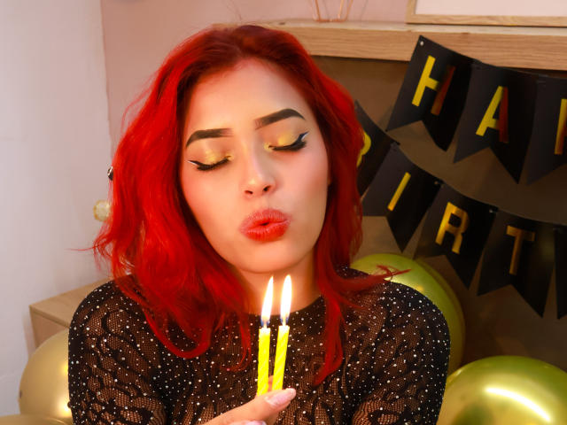 StrawberryX with red hair, closed eyes, and yellow eyeshadow, pouting lips, holding a candle to her nose, wearing a black, sparkly top. Boobs not visible, background ignored.