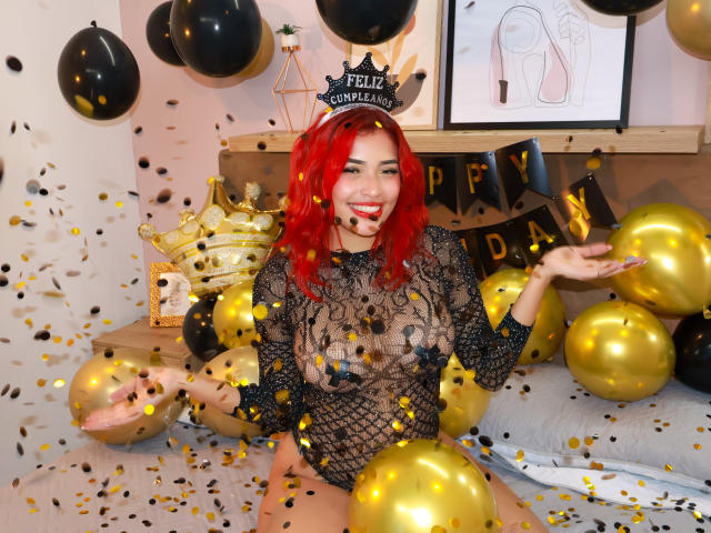 StrawberryX with red hair, wearing a black mesh bodysuit, exposing her tits, surrounded by gold balloons, black confetti, and a "Happy New Year" banner. She's smiling and holding hands.