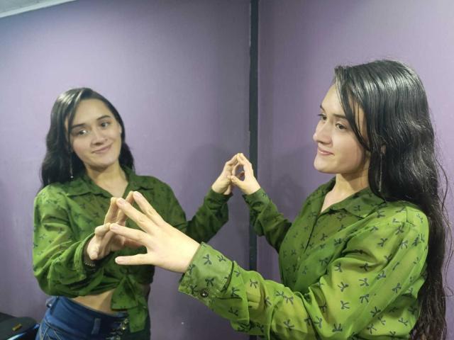 NellyCris with long black hair, green star-patterned shirt, and blue jeans, standing in front of a mirror, smiling while adjusting her hair. Reflects her face and hands.