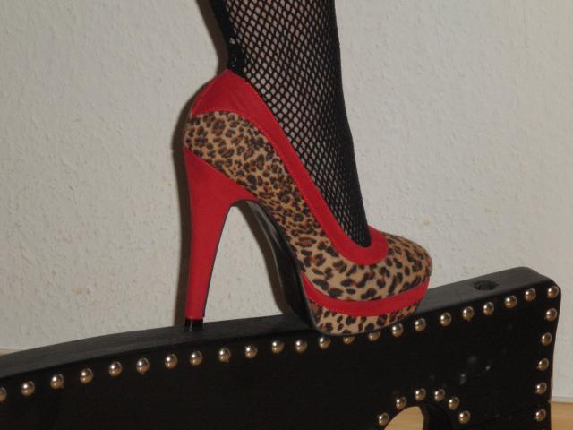 MaitresseGina-hot's foot in a leopard print high heel with red soles and red trim, wearing black fishnet stockings, standing on a black studded leather case.