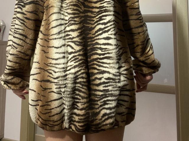 LinaLuchia vestida con un abrigo suave de rayas de tigre, hasta las rodillas, con las manos a los lados, mostrando sus muslos desnudos y una botella a la izquierda en el fondo..