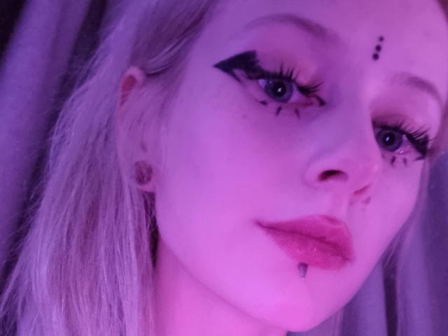 ExcitedBlizzard has pale skin, blue eyes, black winged eyeliner, blonde hair, pink lipstick, and a lip piercing with a small silver ball. Purple light illuminates her face.