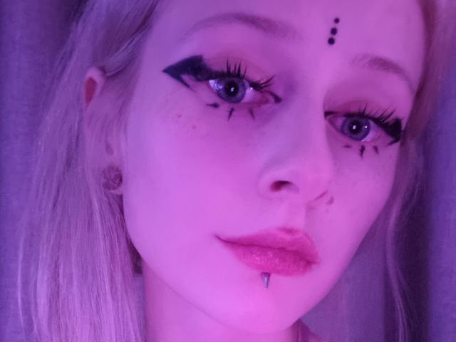 ExcitedBlizzard has purple-tinted skin, long blonde hair, black eyeliner wings, freckles, a nose piercing, a lip piercing with a small ball, and dark purple lipstick.