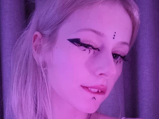 ExcitedBlizzard with pale skin, blonde hair, black eyeliner wings, small black tattoos above eyebrows and chin, and a subtle smile, lit in purple light.