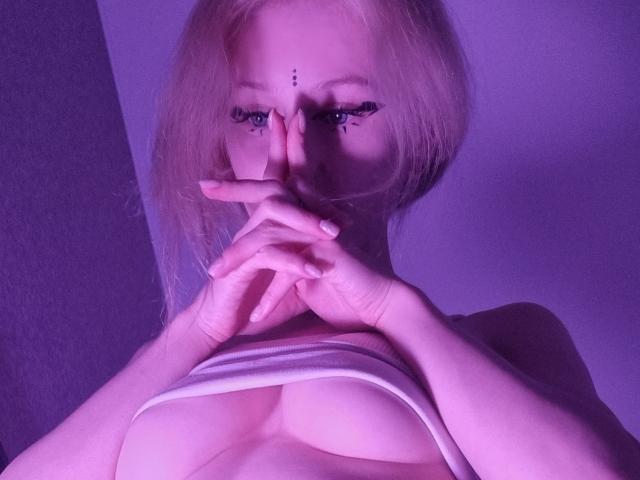 ExcitedBlizzard with blonde hair, eyeliner, and purple lighting, covering mouth with fingers, showing large boobs, white strap barely covering nipples.