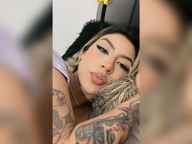 CourtneyLow has tattoos on her arms, pouty pink lips, and heavy eyeliner, lying on a black furry pillow with blonde and black hair, and a yellow flower in background.