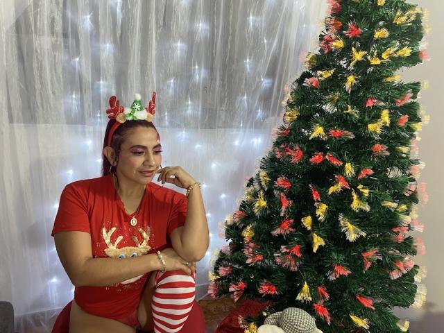 NahelaBigMelonsX, red t-shirt with reindeer, striped thigh-highs, antler headband, sitting next to decorated Christmas tree, white fairy lights, smiling, red panties, curly red hair, hand on chin.