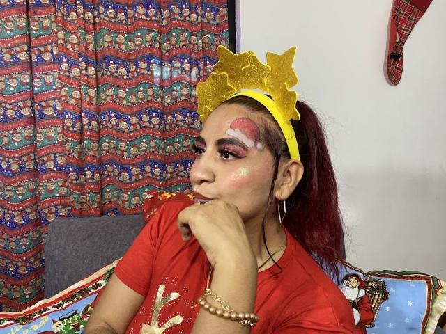 NahelaBigMelonsX with red shirt, yellow headband, face paint, red hair, and gold bangles, pouting with hand on chin, looking left, sitting on colorful Christmas-themed couch.