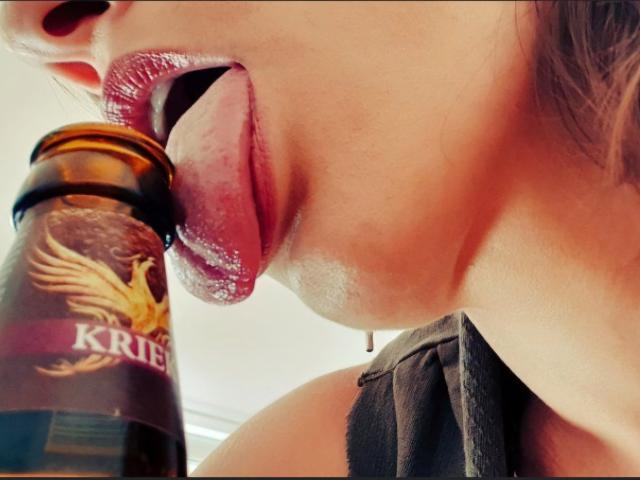 Close-up of JuneLaFrenchie's face with glossy red lips wrapped around a Kruger beer bottle, tongue extended and touching the bottle. Her cheek is flushed, bottle label visible. Sexual tension.