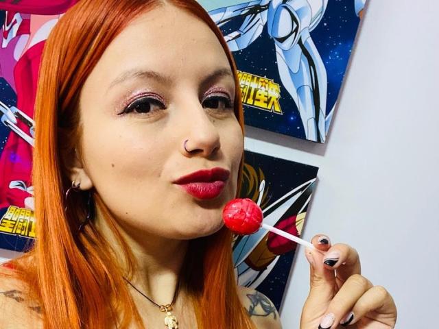 MiiaJonnes with red bra, red panties, and red lipstick, sucking on a red lollipop, has tattoos on her arm, and orange hair, sitting on a black chair.