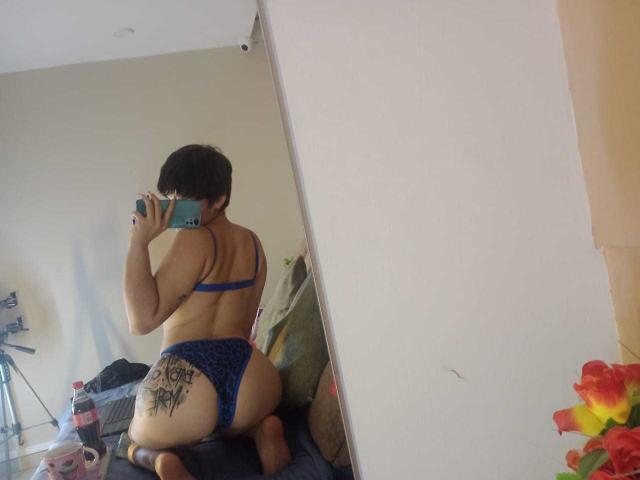 MissTat with short black hair, blue lace bra and panties, tongue out, holding phone, showing tattoo on right ass cheek, kneeling on couch, visible thigh tattoo, tattoos on left thigh.