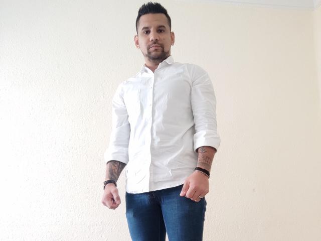 ShadowDiego stands in a white shirt, blue jeans, with tattoos on both arms, short black hair, and a beard, looking serious. He has a slight tan and a muscular build.