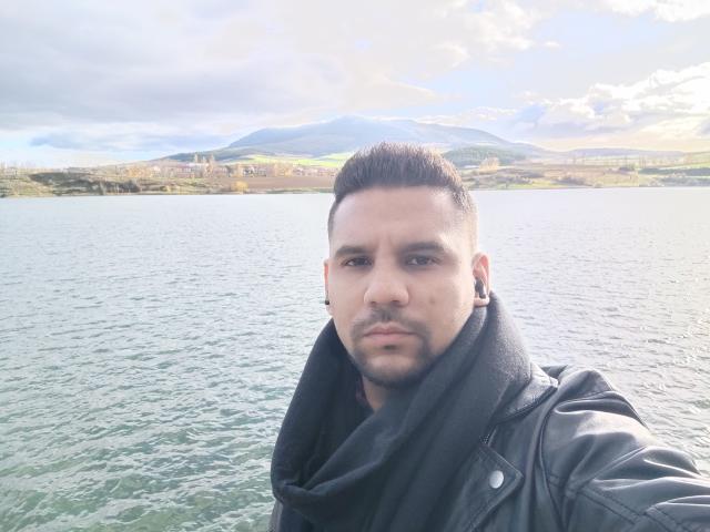 ShadowDiego has a serious face, short brown hair, black leather jacket, black scarf, and black ear gauges. Standing by a greenish-blue lake with mountains in the background.
