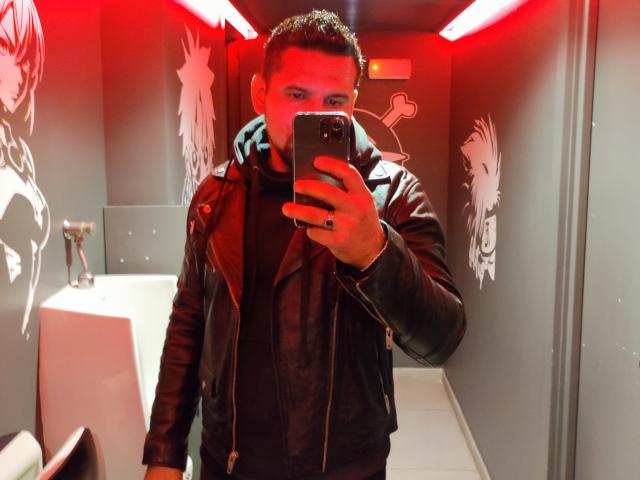 ShadowDiego, taking a mirror selfie in a red-lit bathroom, wearing a black leather jacket, black shirt, and ring, holding a smartphone, with white graffiti on gray walls.