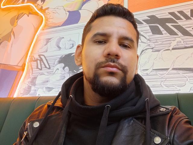 ShadowDiego with a medium brown skin tone, short black hair, and a beard, wearing a black hoodie and leather jacket, has a serious, slightly bored expression.