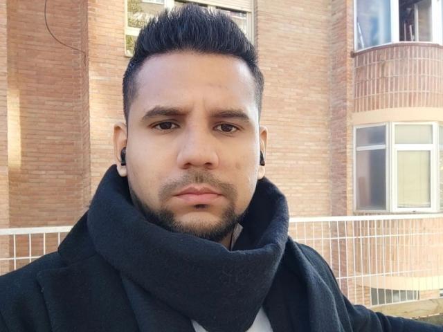 ShadowDiego has a serious face, short black hair, a beard, ear gauges, and wears a thick black scarf with a black coat. He's standing outside in daylight.