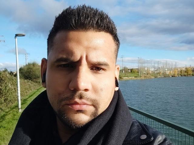 ShadowDiego has a serious face, short dark hair, small goatee, and a black scarf. He's wearing black ear gauges and a leather jacket. His eyes are brown.
